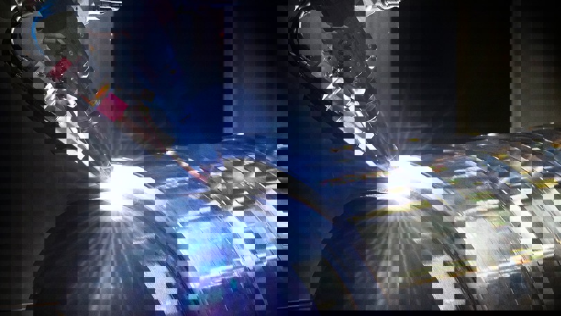 Close up of automated welding torch applying precision weld on cylindrical metal component in a dimly lit industrial setting.