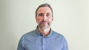 Peter Brock, Senior Account Manager, standing against a plain light background wearing a checkered shirt.