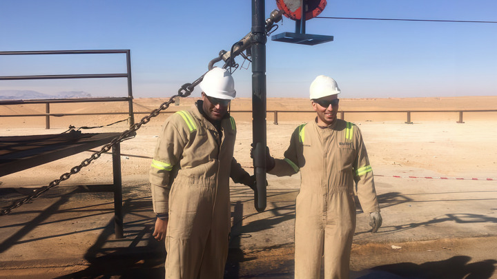 Two oilfield workers in tan coveralls and white helmets stand beside downhole equipment used to recover an irretrievable bridge plug in desert conditions.