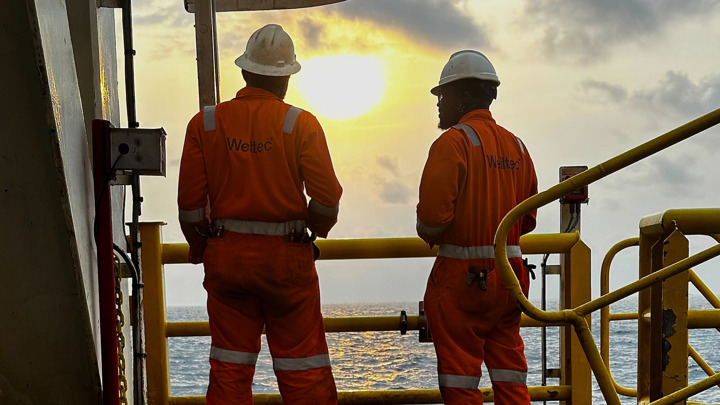 Two Welltec® engineers in orange coveralls observe the sunset from an offshore platform in Angola during a Well Stroker (218) operation.