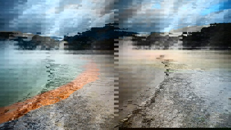 Steam rises from a geothermal hot spring surrounded by colorful mineral deposits and forested hills, illustrating natural geothermal energy.