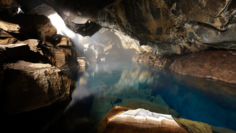 A tranquil cave with a clear blue pool of water reflecting the surrounding rocky walls, illuminated by soft natural light.