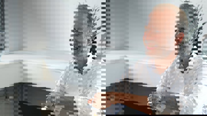 A smiling man in a white shirt sitting at a desk, gesturing with one hand while working on a laptop.