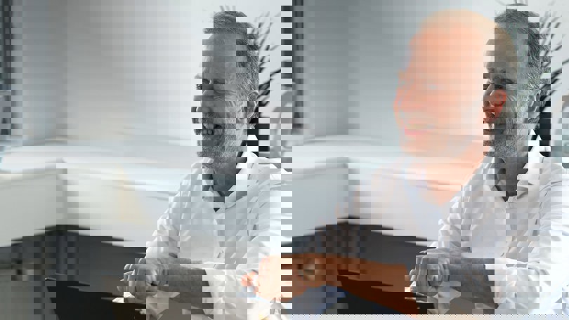 A smiling man in a white shirt sitting at a desk, gesturing with one hand while working on a laptop.