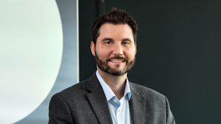 Nick Rakic, Sales Director for Canada, standing against a dark background wearing a gray blazer and smiling.